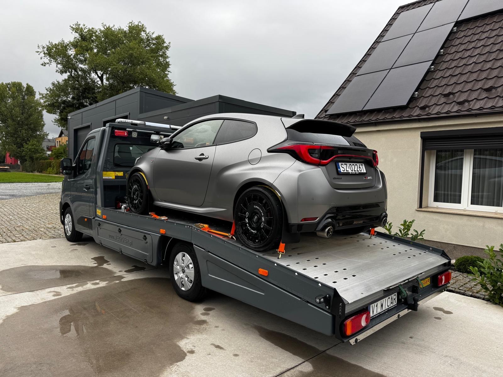 Szara laweta z szarym sportowym autem marki Toyota Yaris GR na platformie, Wrocław. Widoczne panele słoneczne na dachu domu w tle. Auto zabezpieczone pomarańczowymi pasami.