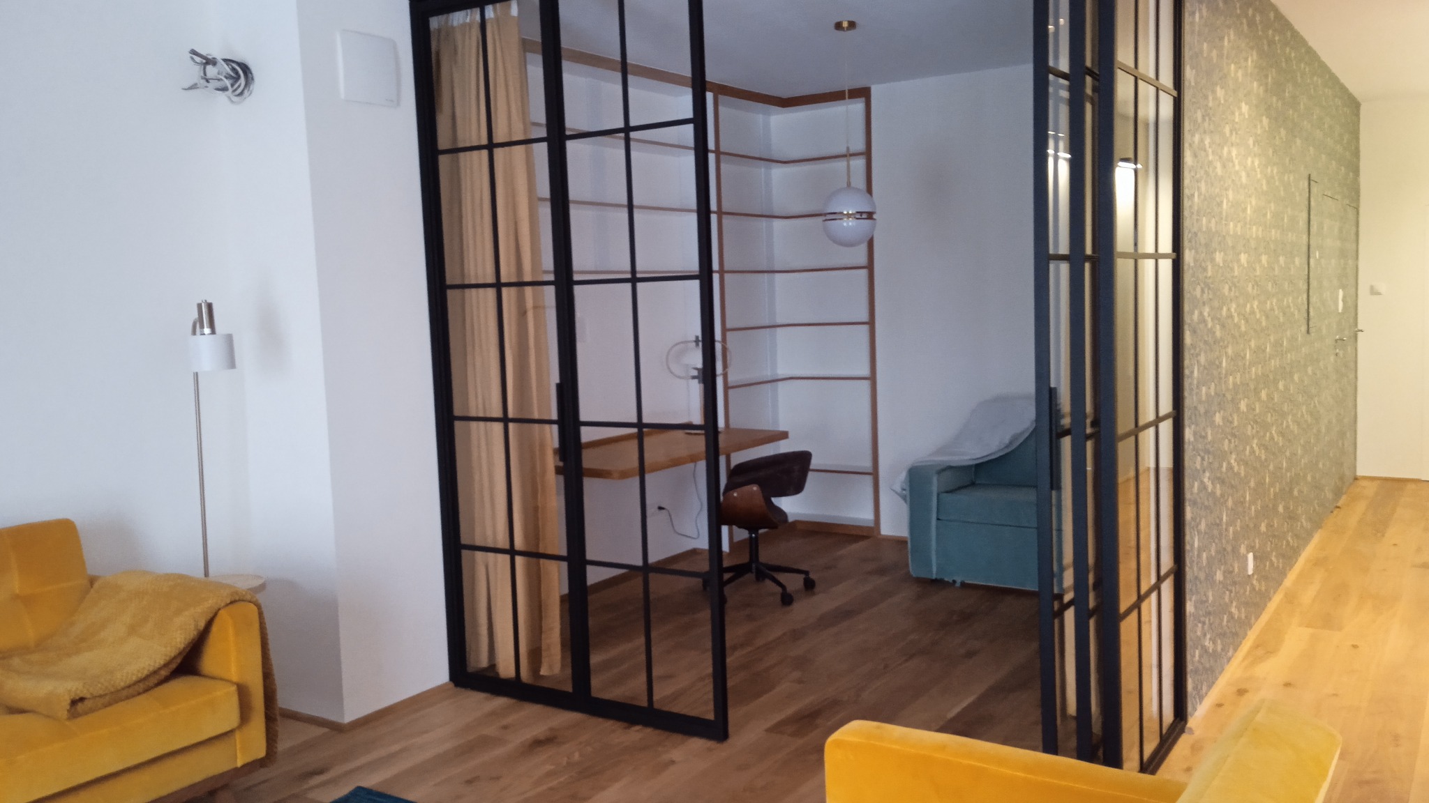 Nowoczesne wnętrze z drewnianą podłogą, ścianą w tapetę i przeszklonymi drzwiami w stylu loft, oddzielającymi pokój z biurkiem i regałem.