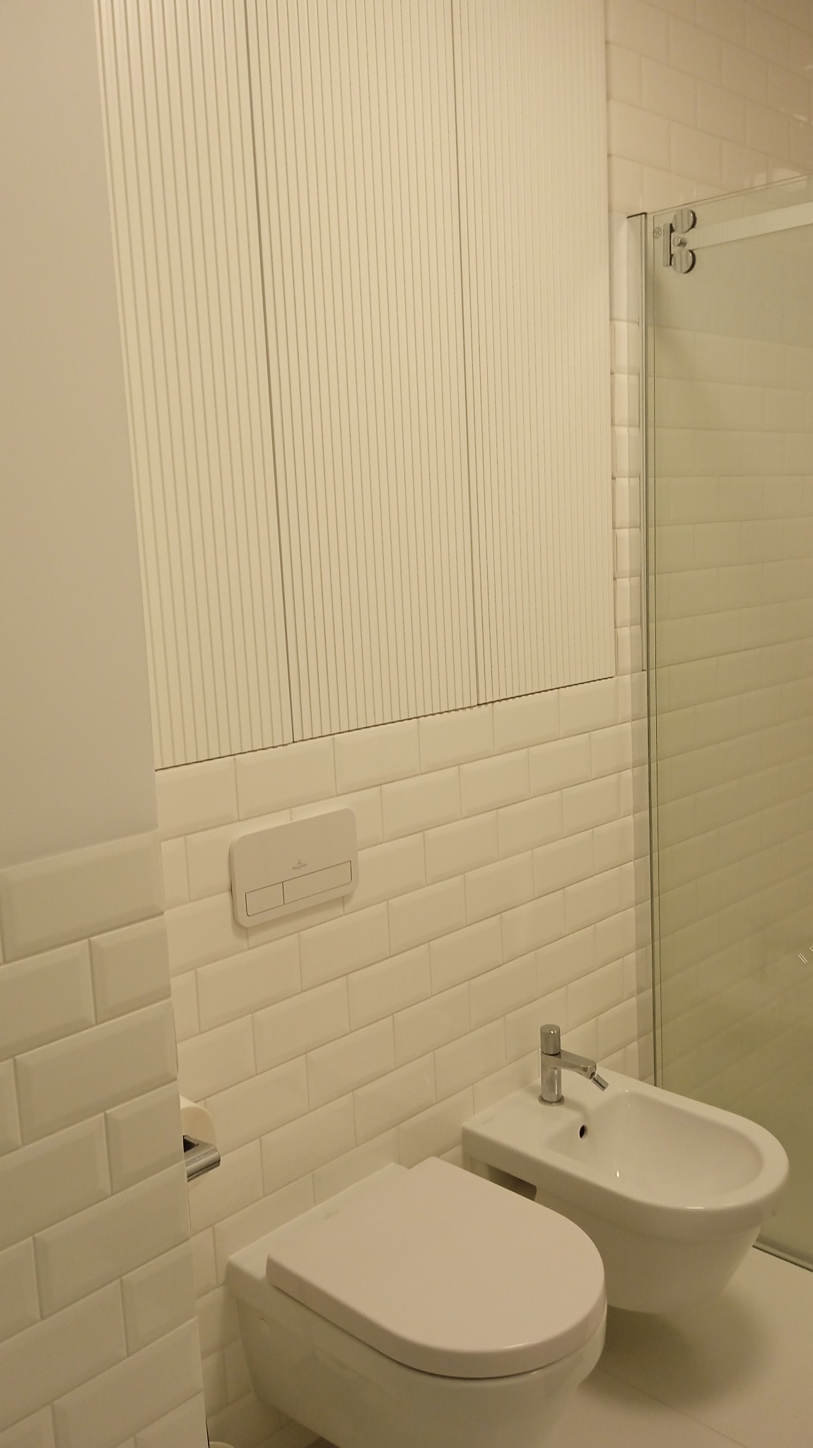 Jasna łazienka z białymi płytkami i armaturą. Widoczne: toaleta, bidet, zabudowa szafkowa z ryflowanymi frontami i szklana kabina prysznicowa. Minimalistyczny design.