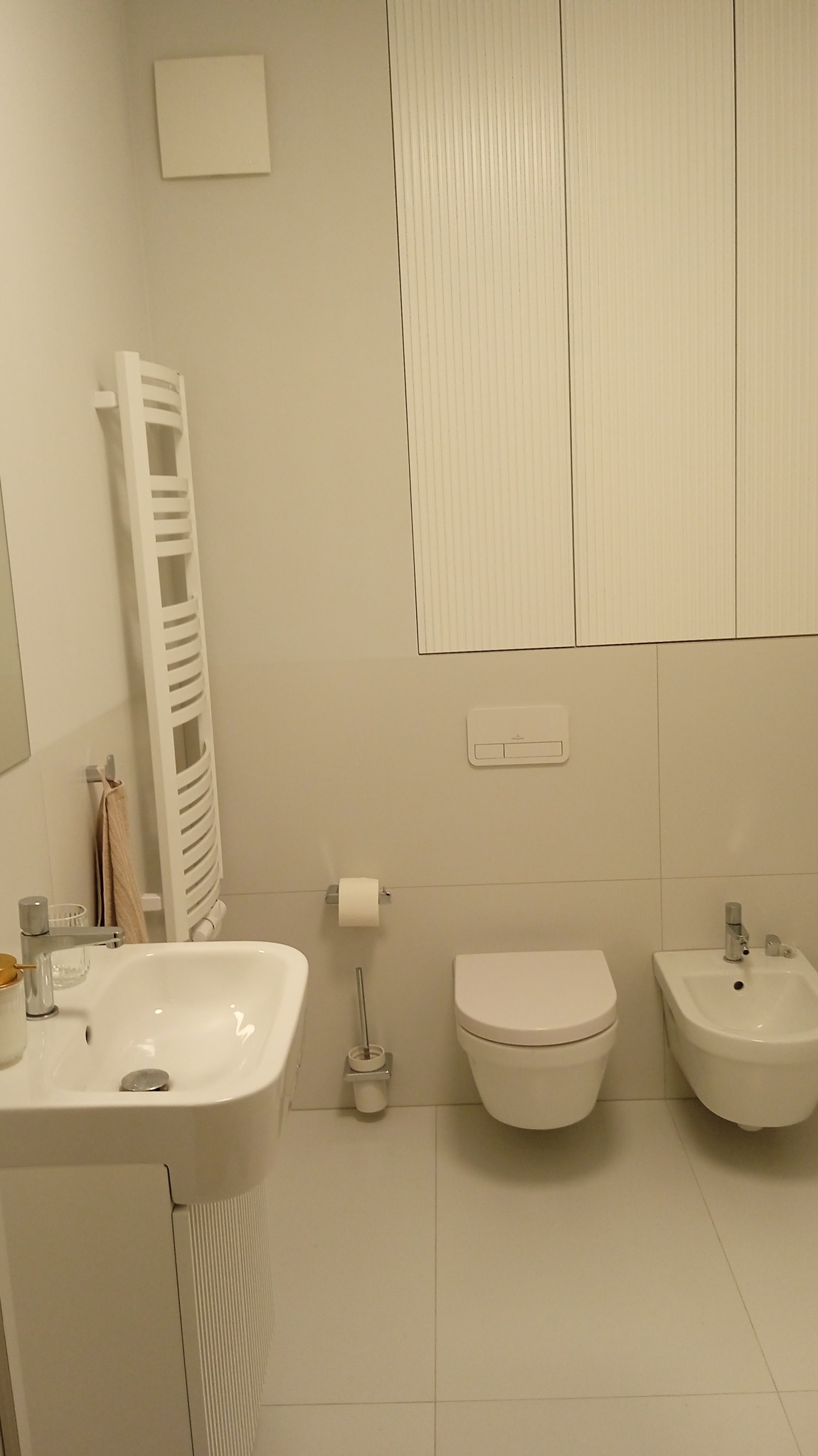 Jasna łazienka z białymi sanitariatami: umywalka na postumencie, toaleta podwieszana i bidet. Na ścianie grzejnik drabinkowy i szafki z lamelami.