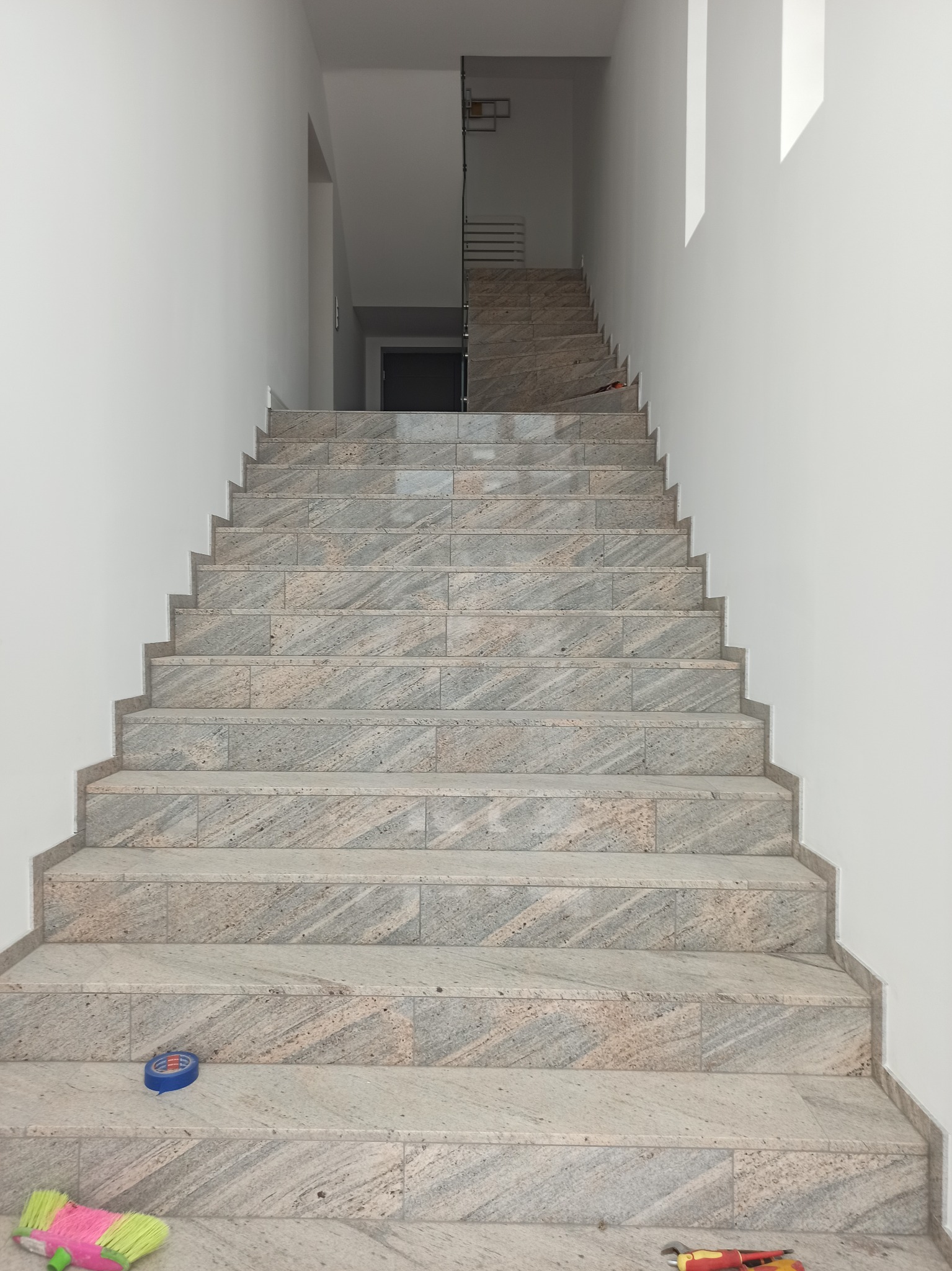 Wykończone schody wewnątrz budynku, wyłożone płytkami imitującymi kamień. Widoczne narzędzia glazurnicze na stopniach. Ujęcie od dołu, perspektywa w górę.