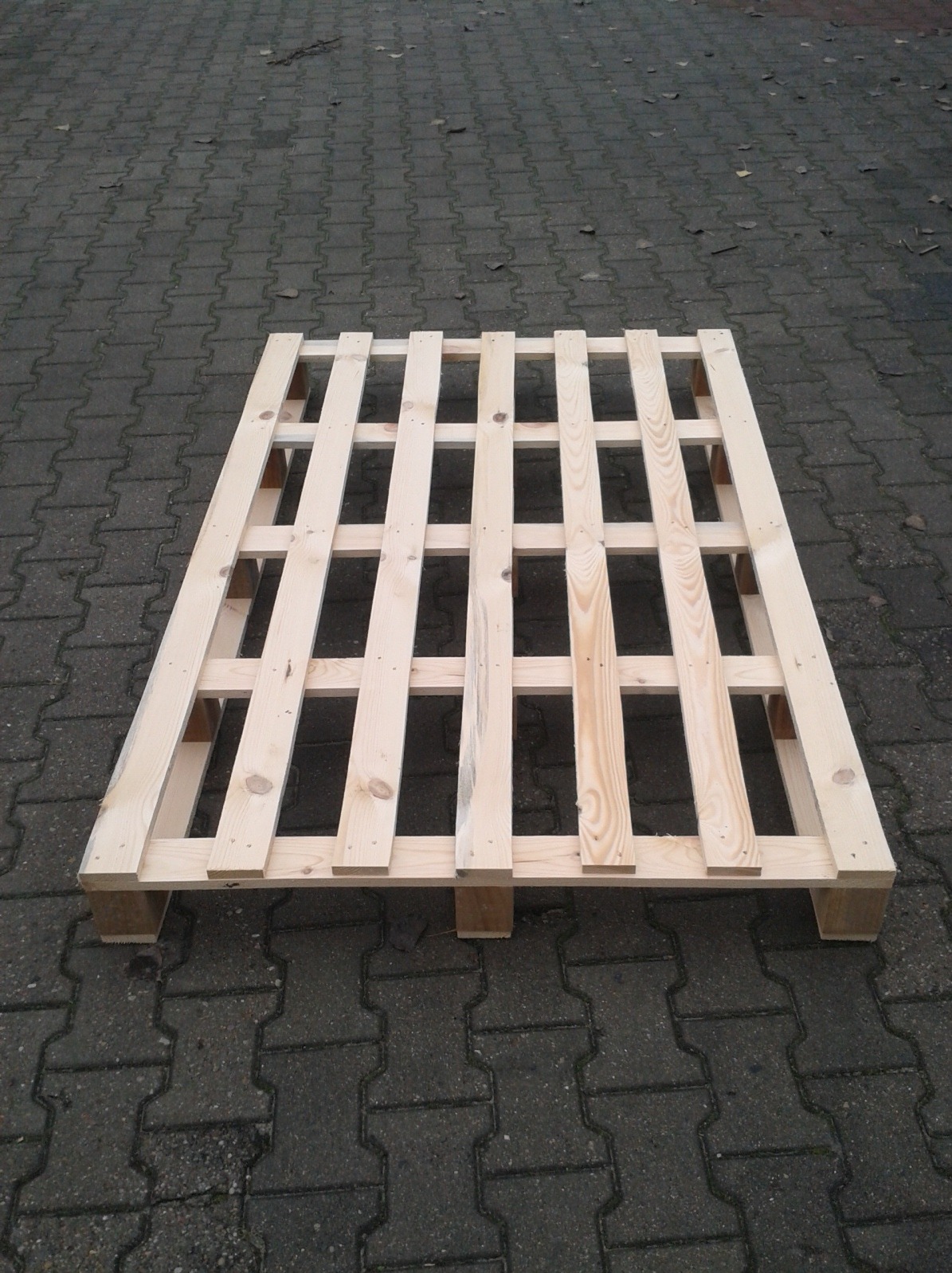 Jasna, drewniana paleta na szarym bruku. Widok z góry, ukazujący konstrukcję kratową palety i jej solidne wykonanie. Tło stanowią liście na ziemi.