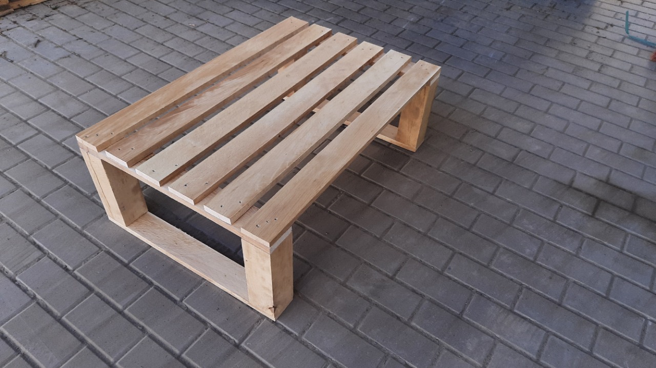 Niski stolik kawowy z jasnego drewna paletowego, ustawiony na szarej kostce brukowej. Widoczne gwoździe łączące elementy. Minimalistyczny design.