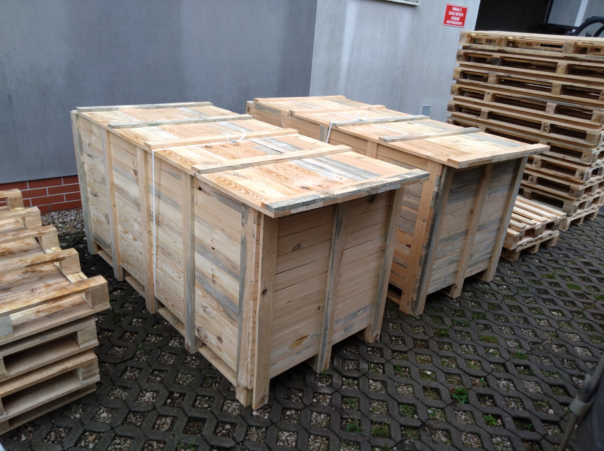 Dwa drewniane kontenery na paletach, zabezpieczone taśmami, stoją na betonowej kostce. W tle ściana budynku i stos palet.