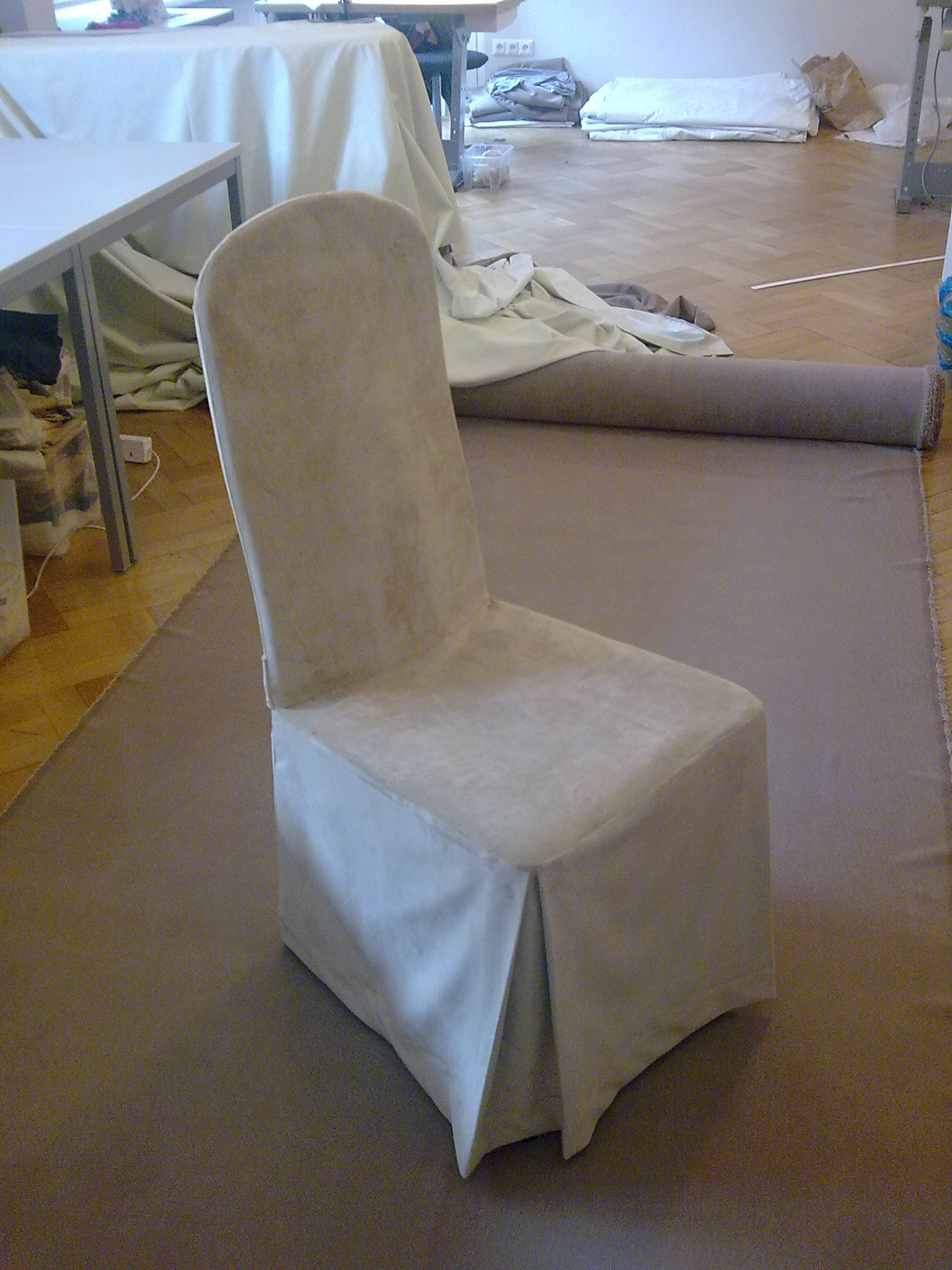 Krzesło w pokrowcu z falbaną, leżące na rozłożonym dywanie, w tle materiały i stół krawiecki.