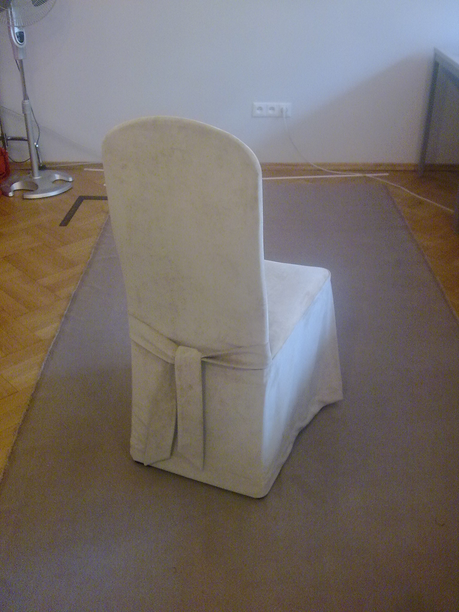 Krzesło z kremowym pokrowcem wiązanym z tyłu, stojące na szarym dywanie, widoczne gniazdko elektryczne i wentylator w tle.