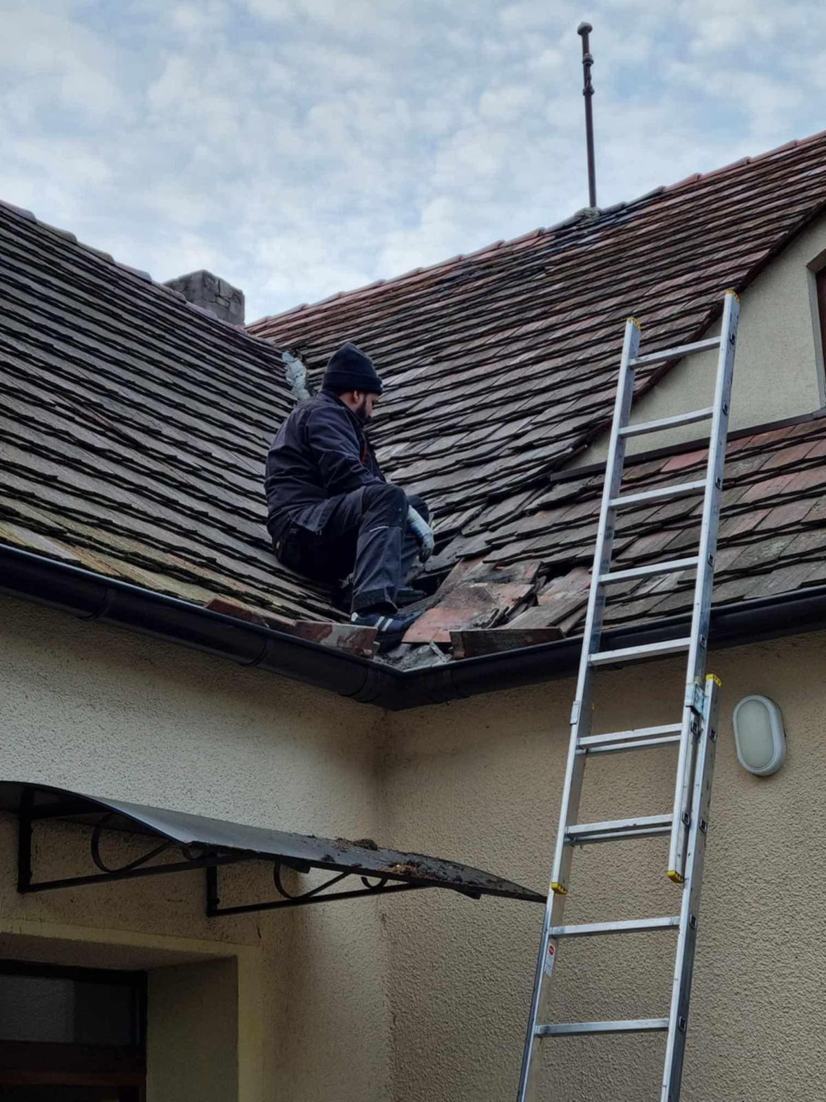 Dekarz w czarnym ubraniu siedzi na dachu z uszkodzonymi dachówkami, obok drabina. Widoczne narzędzia i fragment elewacji budynku. Praca na wysokości.