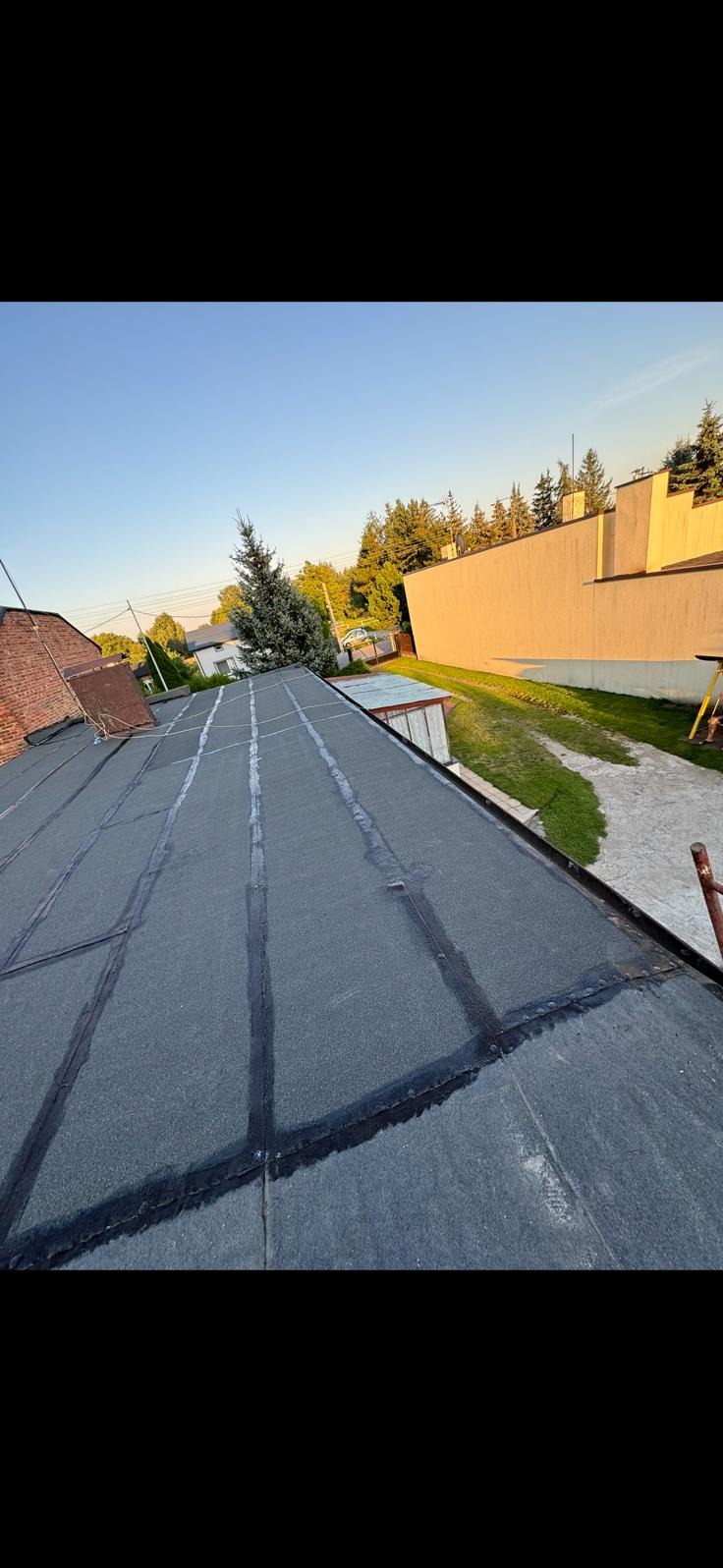 Dach pokryty papą termozgrzewalną, widoczne łączenia. W tle budynki i drzewa w letnim słońcu. Ujęcie z góry, lekko z boku.