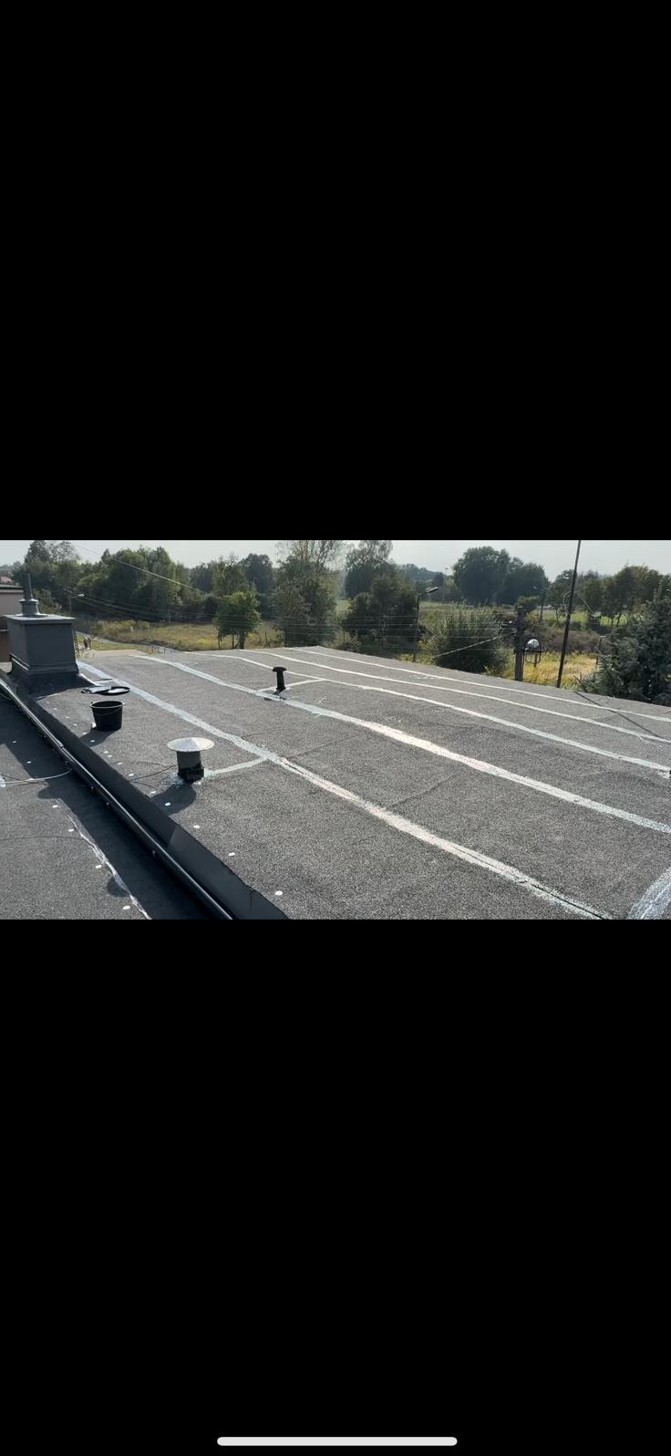 Płaski dach pokryty papą z widocznymi pasami uszczelniającymi, wentylacja i wiadro na tle zieleni. Widok z góry na wykończony dach.