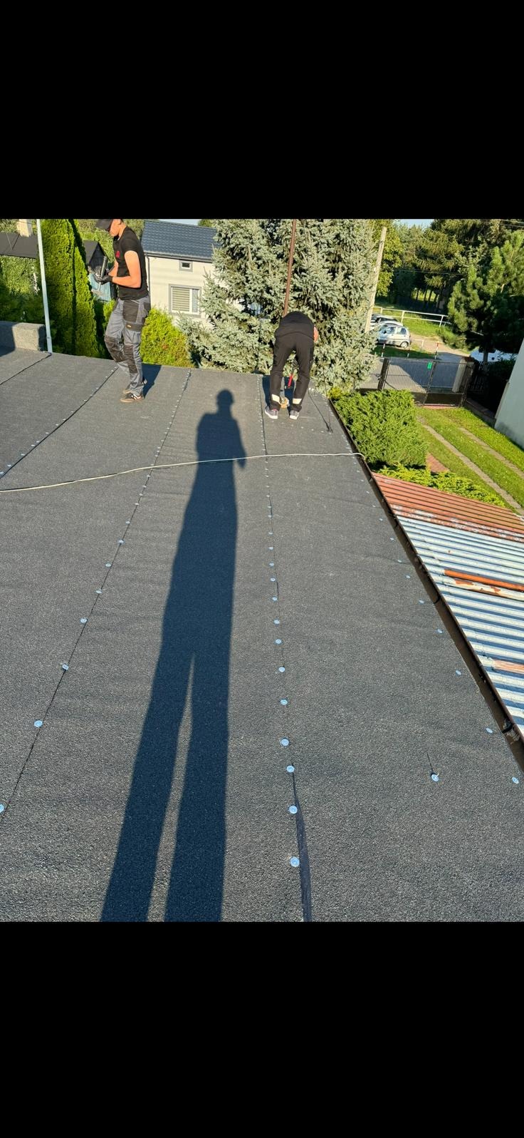 Praca na dachu pokrytym papą. Widoczne mocowania, cień osoby i fragmenty otoczenia: drzewa i budynki. Dwóch dekarzy w trakcie pracy.