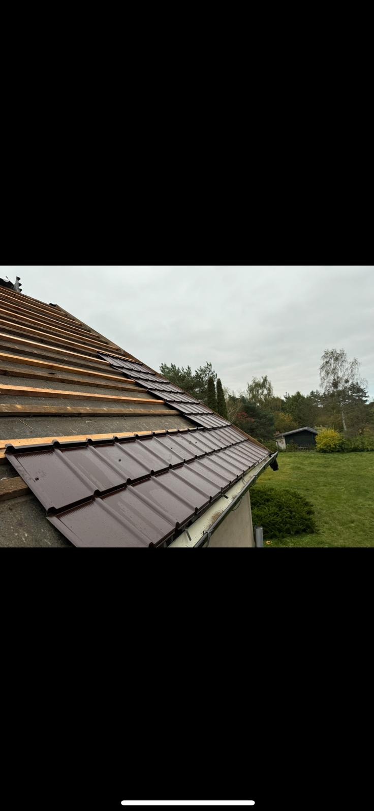 Układanie brązowej blachodachówki na dachu domu jednorodzinnego. Widoczne drewniane łaty i rynna. Częściowo pokryty dach na tle zieleni i pochmurnego nieba.