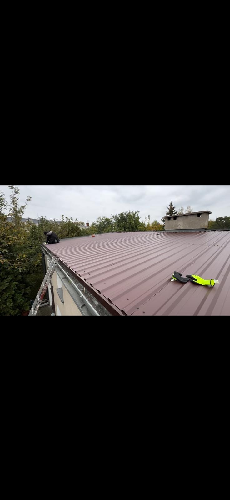 Ciemnoczerwony blaszany dach z rynnami, na którym siedzi dekarz. Na dachu leżą też żółte rękawice robocze. Widoczne drzewa i komin w tle.