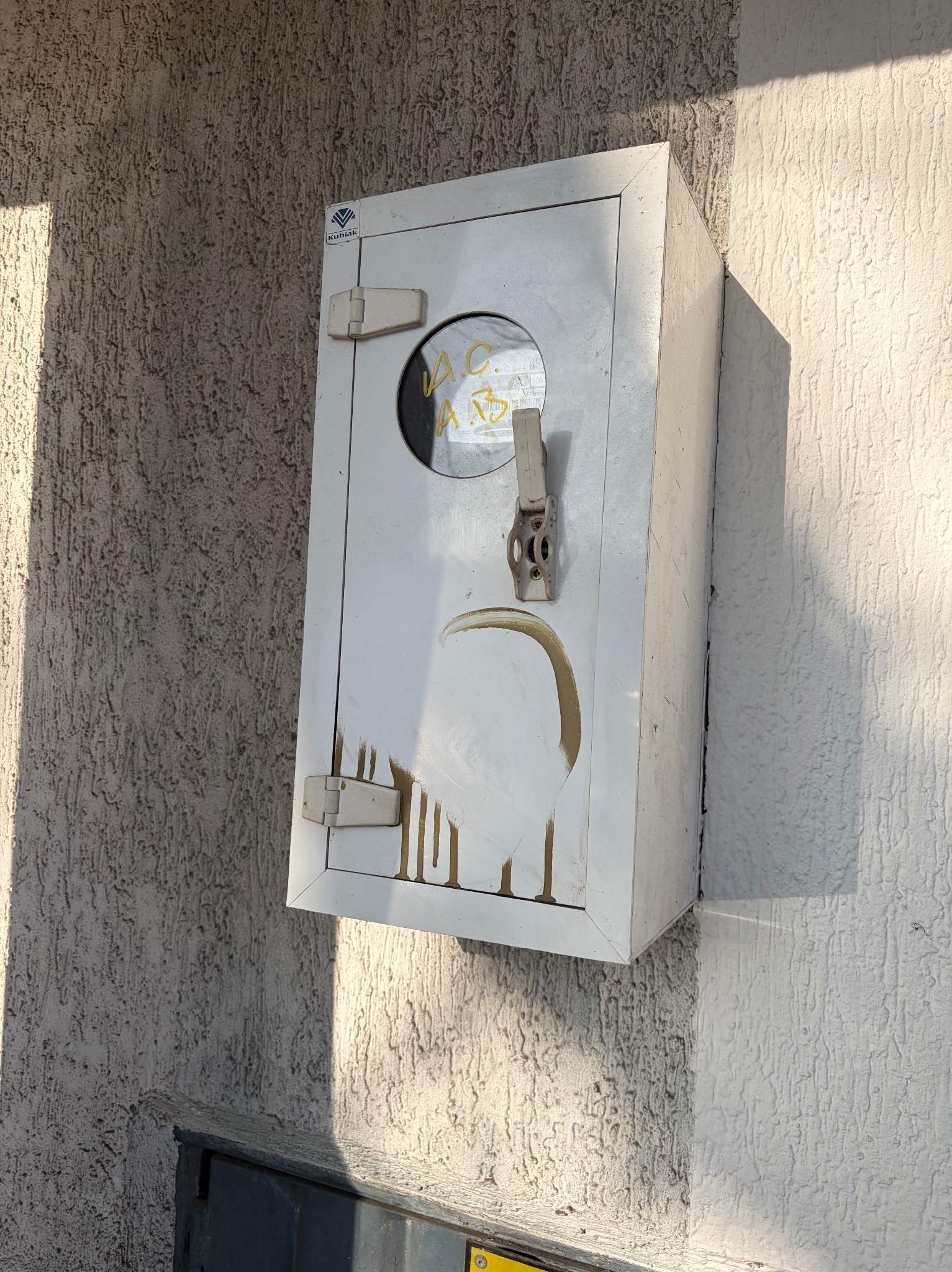 Biała skrzynka na ścianie z fakturą baranka, z widocznymi śladami po czyszczeniu. Okrągłe okienko z żółtymi napisami. Oświetlenie słoneczne z lewej strony.