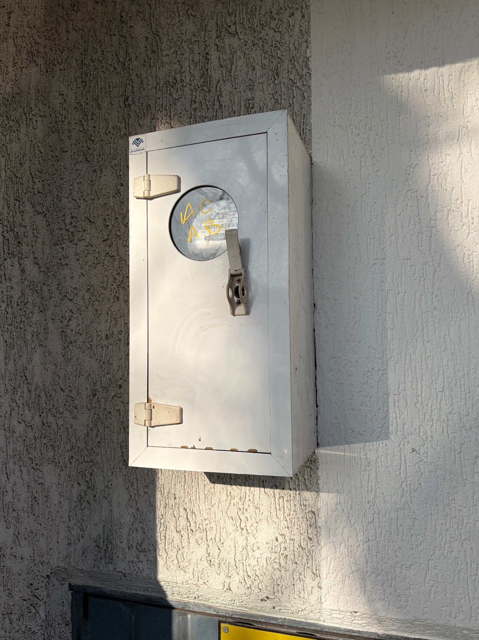 Metalowa skrzynka z licznikiem na elewacji budynku o nierównej fakturze, oświetlona bocznym światłem słonecznym, z widocznym logo producenta.