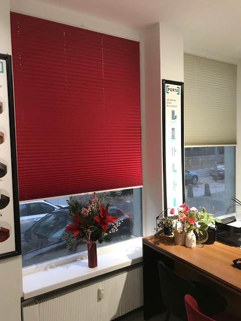 Czerwona, plisowana osłona okienna w pomieszczeniu z dekoracjami świątecznymi na parapecie. W tle widoczne okno z jasną osłoną i fragment biurka.