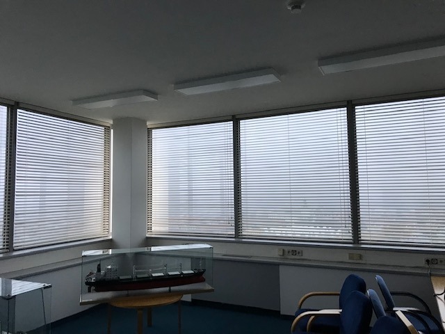 Jasne biuro z dużymi oknami, na których zamontowano poziome, aluminiowe osłony. W tle widok na miasto. W pomieszczeniu model statku w szklanej gablocie.