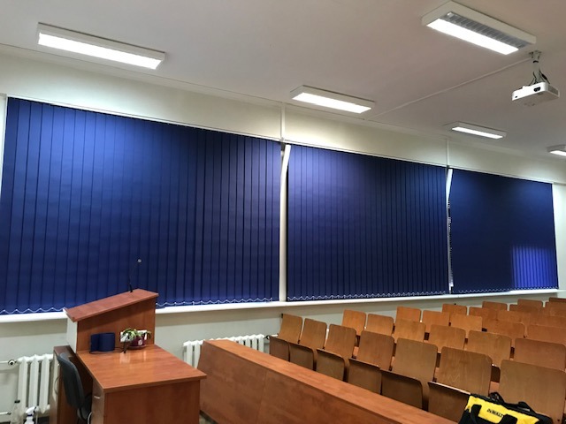 Niebieskie pionowe osłony okienne w sali wykładowej, widoczne krzesła i mównica. Oświetlenie sufitowe i projektor. Jedna osłona częściowo rozsunięta.