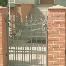 Kuta brama wejściowa z ozdobnymi detalami, osadzona między ceglanymi słupkami, prowadząca do domu z widocznymi schodami.