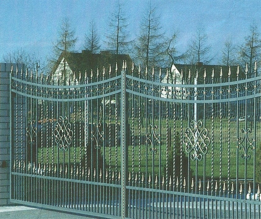 Otwarta, metalowa brama w kolorze szarym z dekoracyjnymi elementami w kolorze złotym, prowadząca do posesji z domem i zielenią w tle.