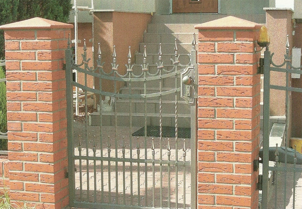 Kuta brama wejściowa z ozdobnymi detalami, osadzona między ceglanymi słupkami, prowadząca do domu z widocznymi schodami.