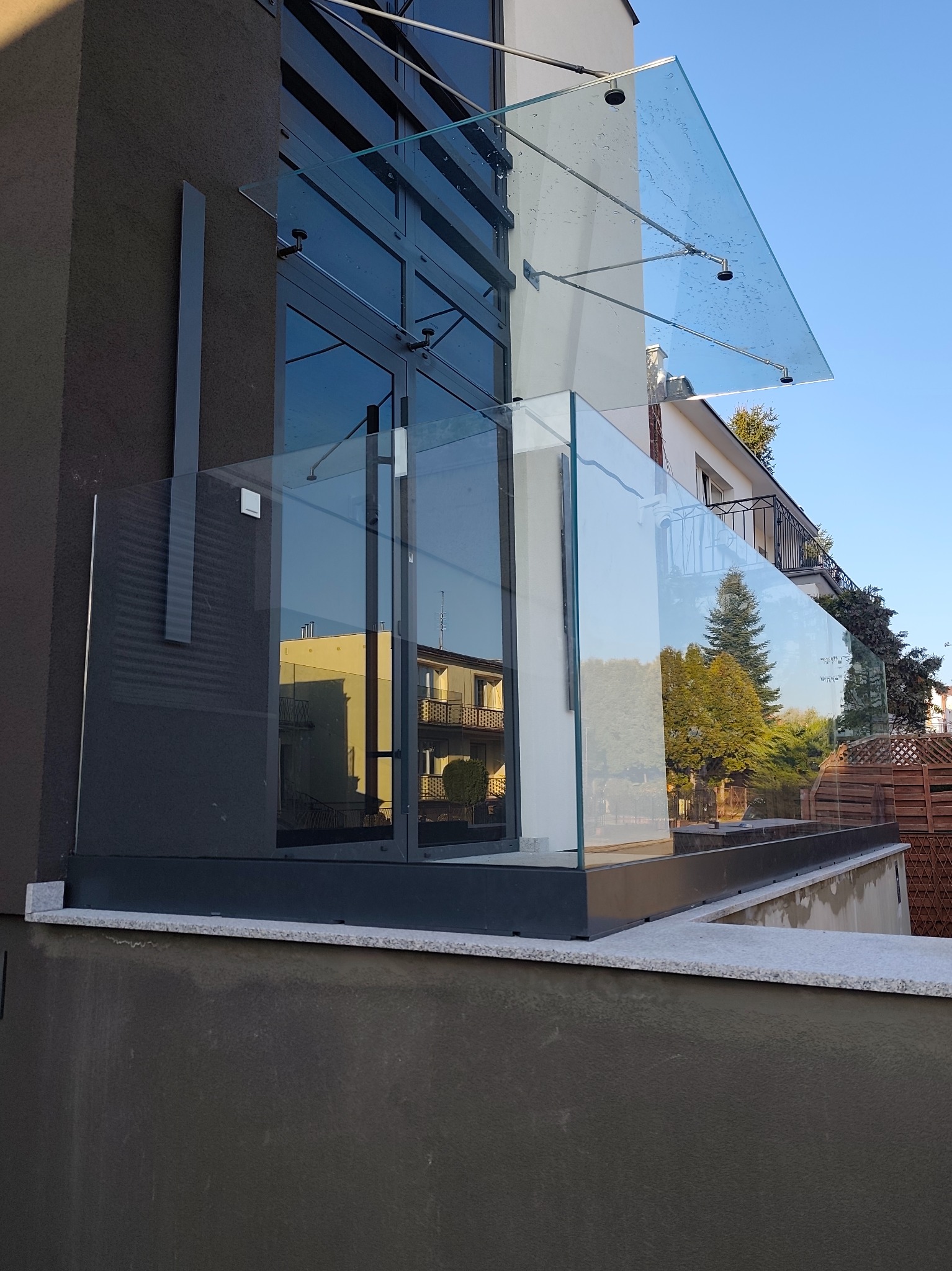 Nowoczesna szklana balustrada balkonowa z daszkiem, mocowana do elewacji budynku. Widoczne detale mocowań i refleksy otoczenia w szkle.