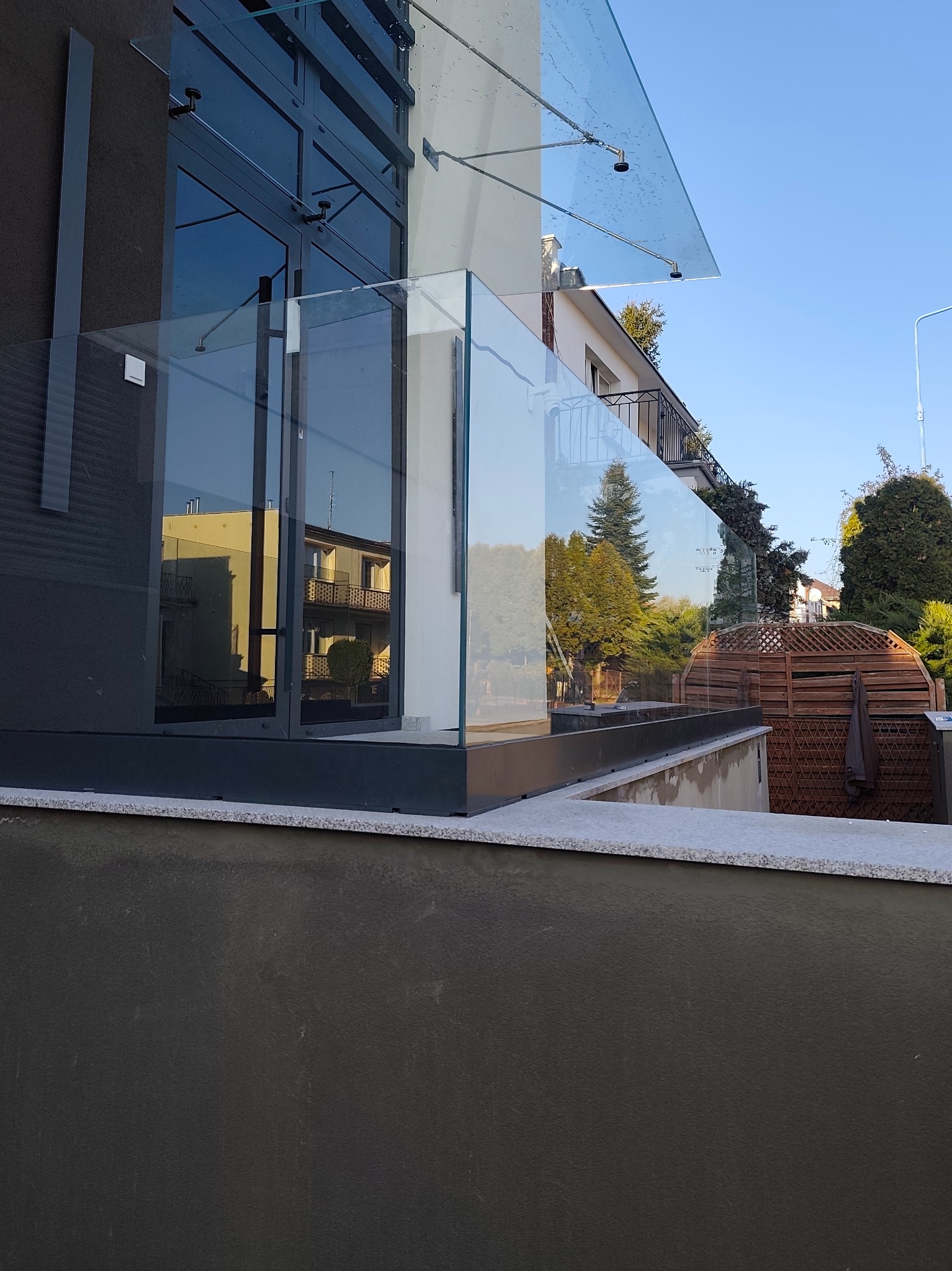 Szklana balustrada balkonowa z widocznym odbiciem budynku w tafli szkła. Nowoczesny design z elementami metalowymi i drewnianą osłoną w tle.