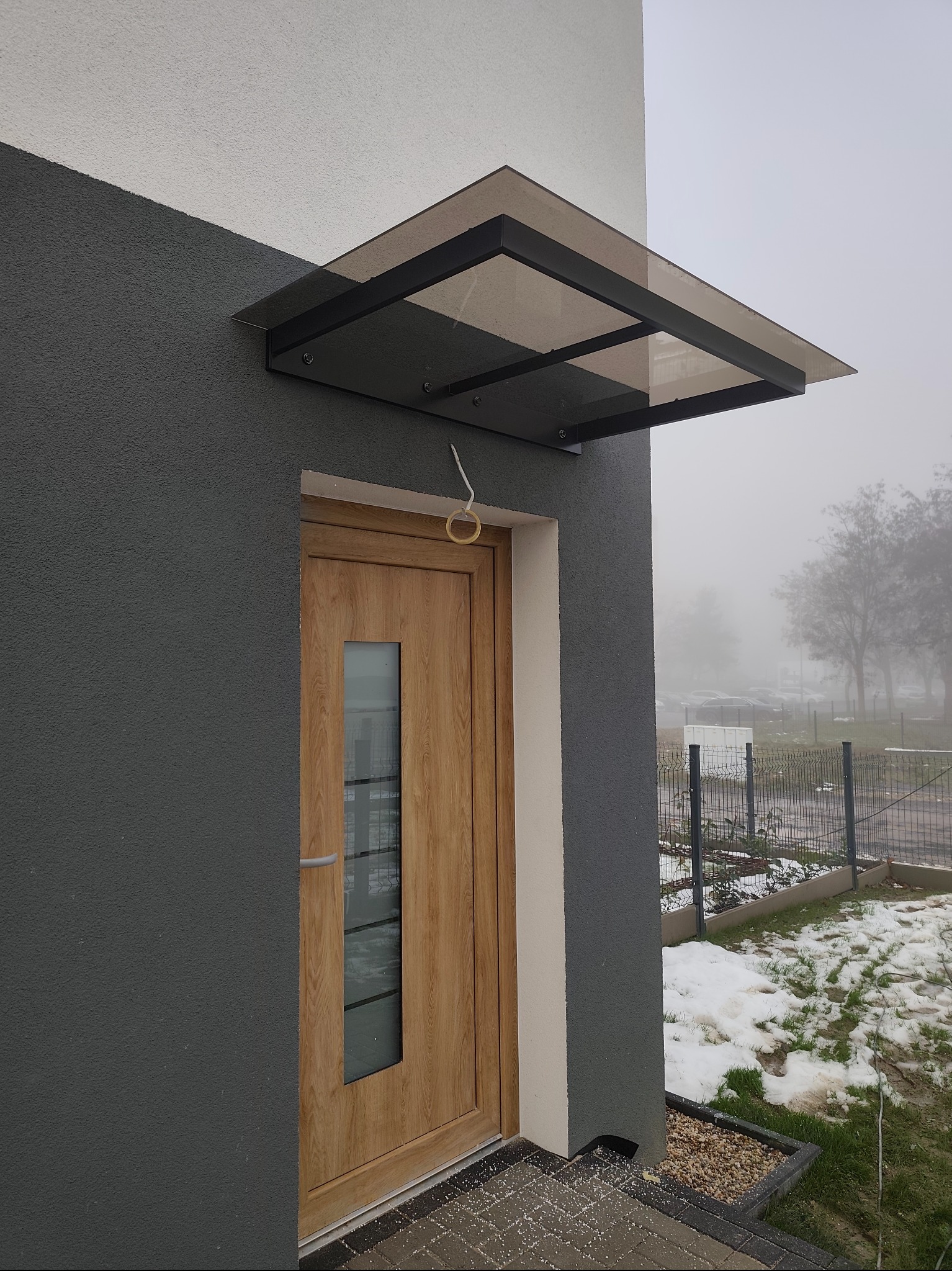 Nowoczesne zadaszenie nad drzwiami wejściowymi z czarną ramą i przydymionym szkłem, zamontowane na elewacji budynku w mglisty dzień.