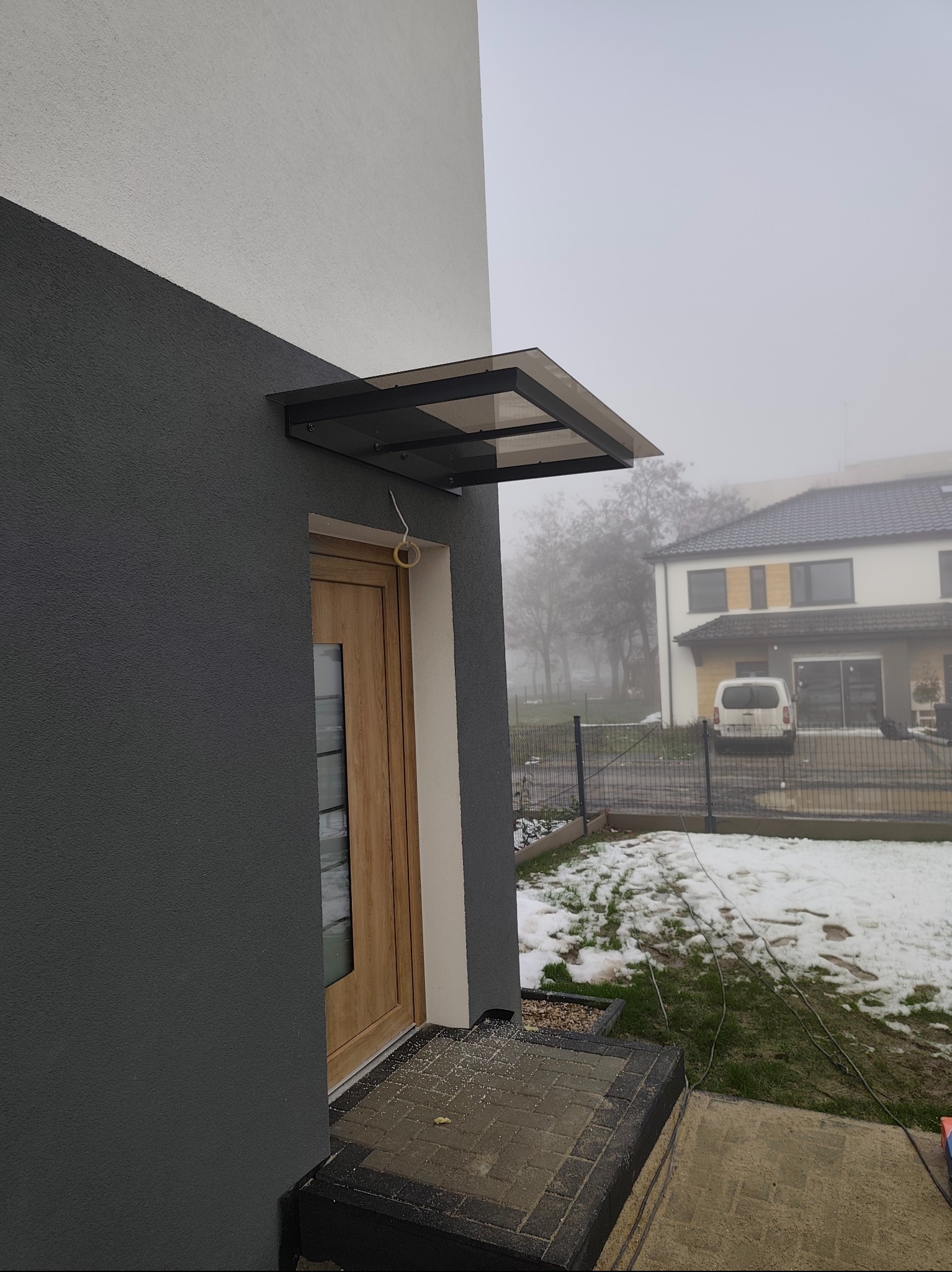 Nowoczesne zadaszenie nad drzwiami wejściowymi, wykonane z metalu i przezroczystego tworzywa. Widoczne ślady wiercenia na kostce brukowej. Dom w tle, pochmurny dzień.