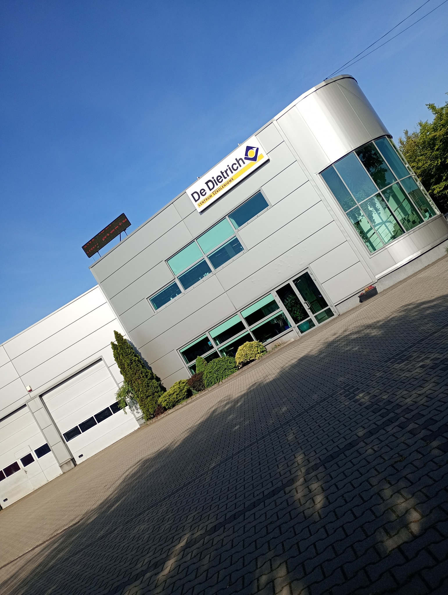 Nowoczesny budynek z logo De Dietrich pod błękitnym niebem, widziany pod kątem. Widoczne okna, drzwi i fragment parkingu z kostki brukowej. Budynek otoczony zielenią.