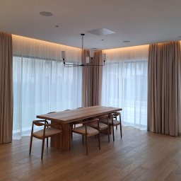 Deco Inspiration Joanna Michalak - Eleganckie, beżowe zasłony z delikatnej tkaniny w przestronnym salonie z drewnianym stołem i krzesłami. Subtelne oświetlenie LED podkreśla karnisze.