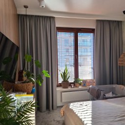 Deco Inspiration Joanna Michalak - Szare zasłony na karniszu, drewniane żaluzje w oknie, rośliny doniczkowe na parapecie, łóżeczko dziecięce w pokoju z delikatnym światłem słonecznym.