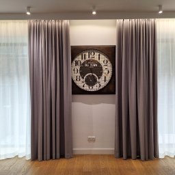Deco Inspiration Joanna Michalak - Eleganckie, długie zasłony w odcieniach szarości i białe firany, symetrycznie ułożone po bokach ściany z dekoracyjnym zegarem. Parkiet na podłodze.