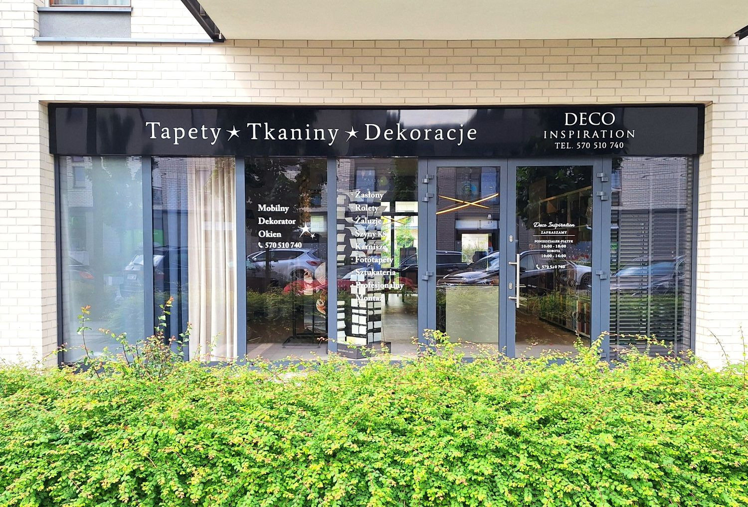 Elewacja lokalu 'Deco Inspiration' z ofertą tapet, tkanin i dekoracji okiennych. Widoczne zasłony, rolety i żaluzje. Na szybie wypisane usługi i dane kontaktowe.