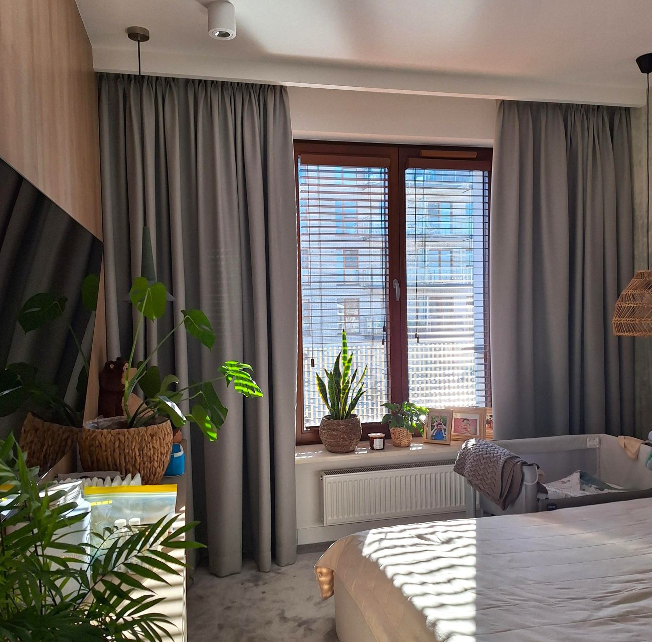Szare zasłony na karniszu, drewniane żaluzje w oknie, rośliny doniczkowe na parapecie, łóżeczko dziecięce w pokoju z delikatnym światłem słonecznym.
