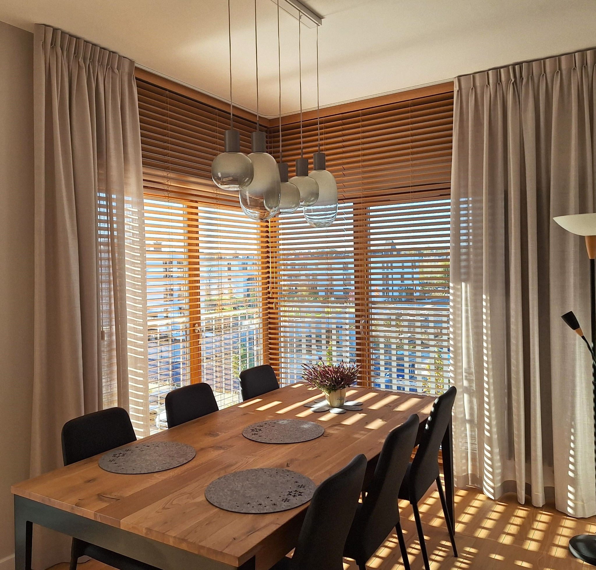Jadalnia z drewnianymi żaluzjami, beżowymi zasłonami i lampami wiszącymi. Promienie słoneczne wpadają przez okno, oświetlając drewniany stół i czarne krzesła.