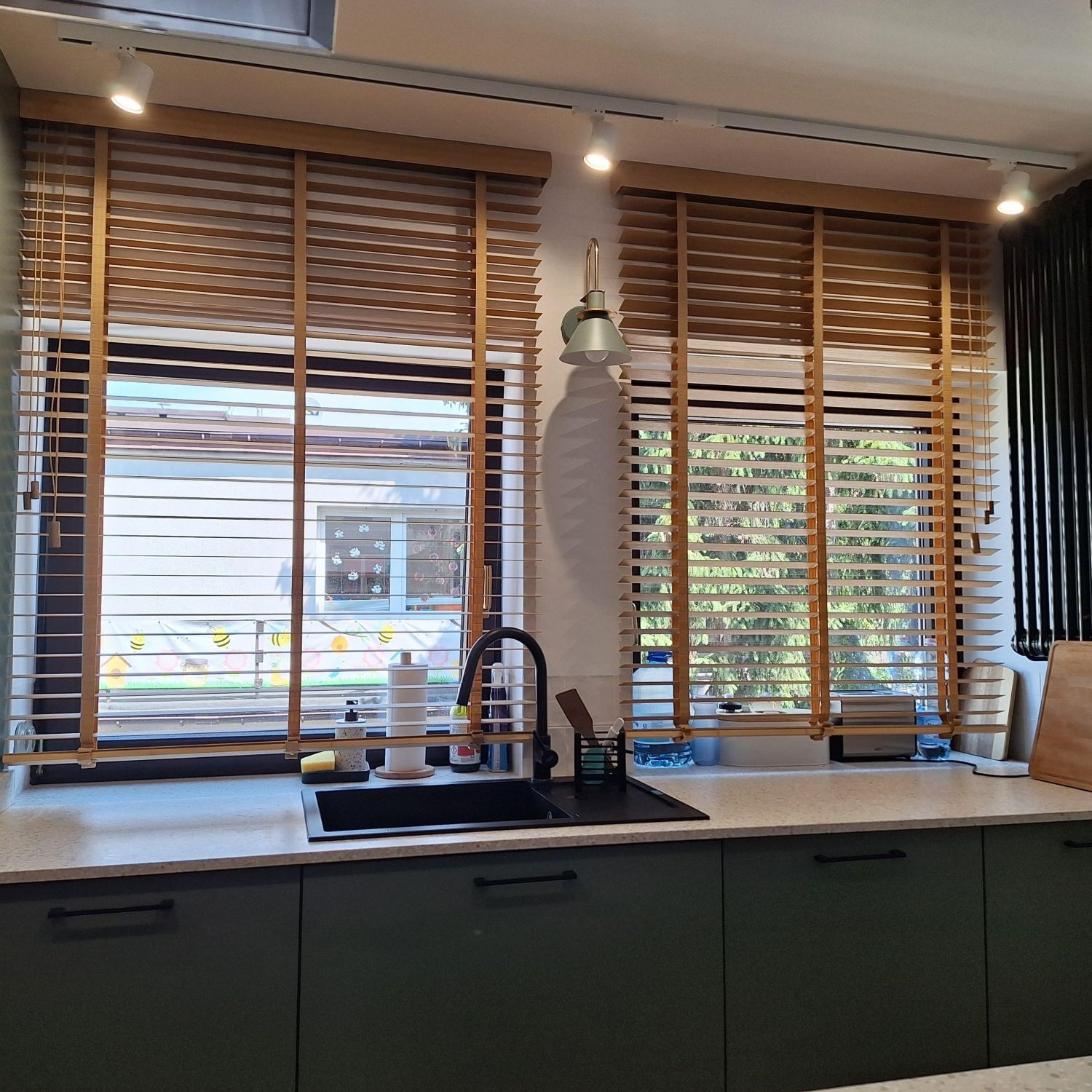 Kuchnia z drewnianymi żaluzjami w oknach, przez które widać ogród. Blat z czarnym zlewem i ciemnozielone szafki. Oświetlenie punktowe.