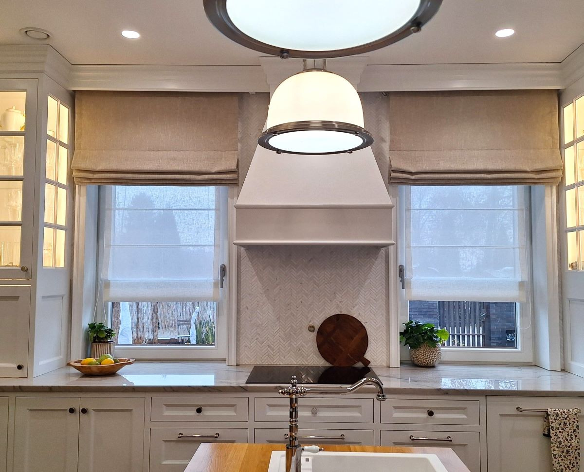 Elegancka kuchnia z beżowymi roletami rzymskimi w oknach, marmurowym blatem i białymi szafkami. Centralnie umieszczony okap i lampa sufitowa.