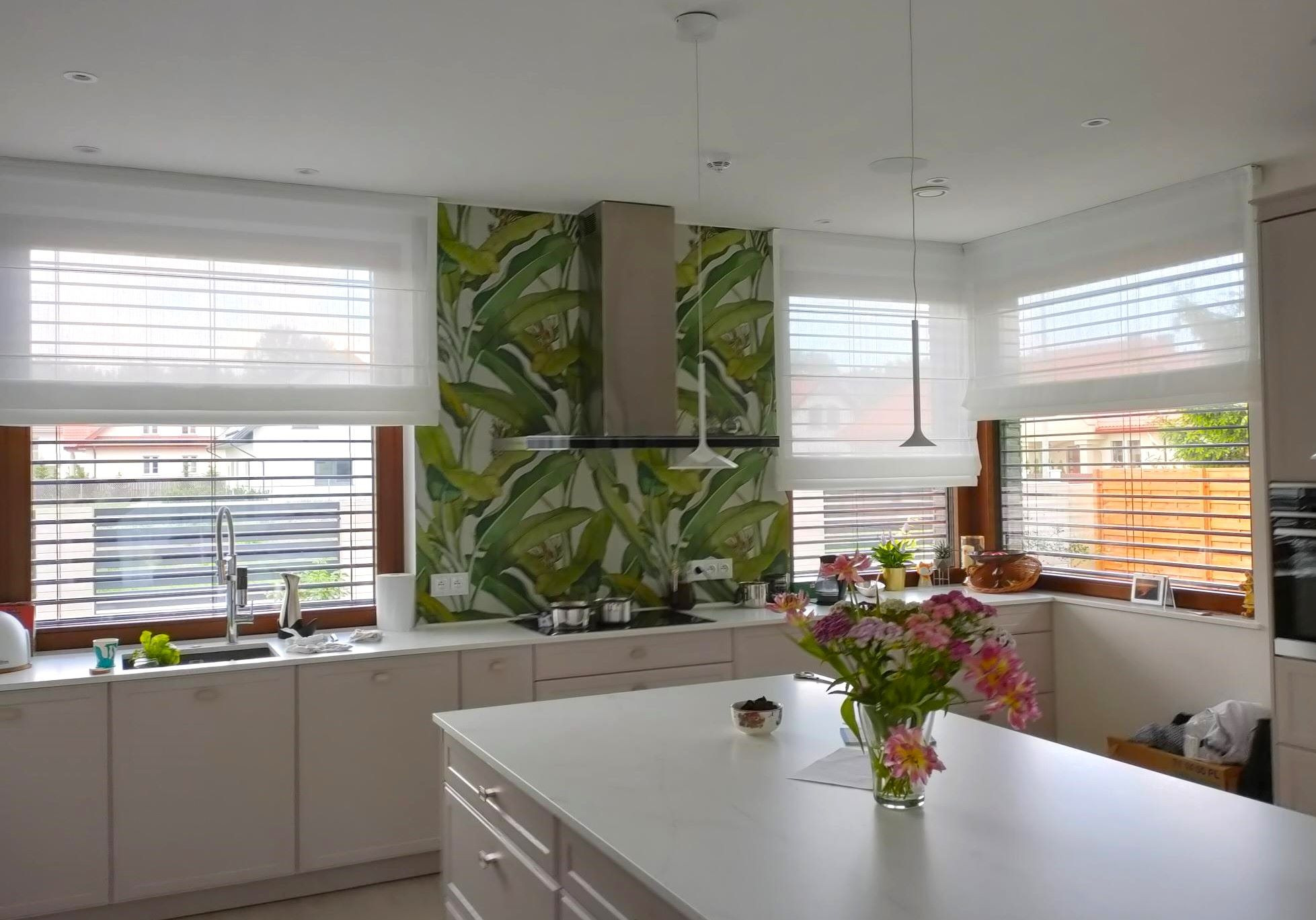 Jasna kuchnia z białymi roletami rzymskimi w oknach i tapetą z motywem liści bananowca nad blatem. Minimalistyczne lampy wiszące.