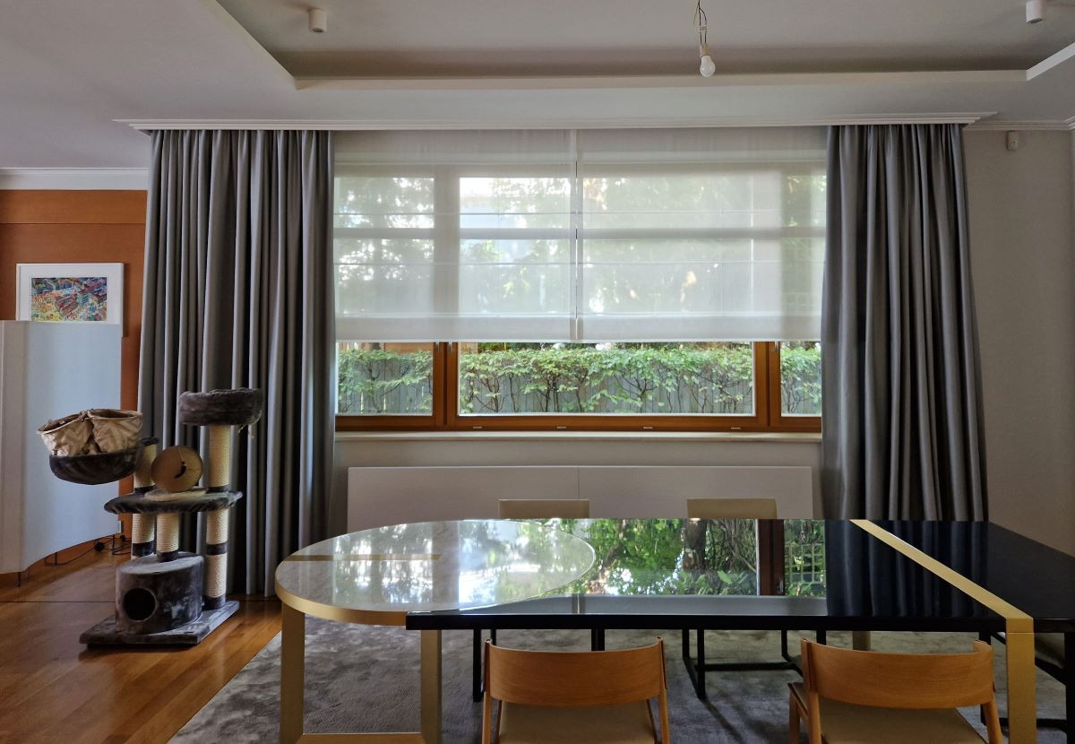 Eleganckie szare zasłony i jasne rolety rzymskie w oknie, widok na zieleń zza okna, nowoczesny stół w jadalni, drapak dla kota w tle.