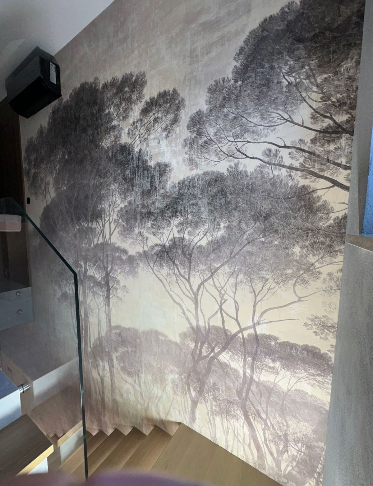 Tapeta z motywem leśnym zdobi ścianę przy schodach w Warszawie. Delikatne, stonowane barwy tworzą spokojną atmosferę. Widoczny fragment szklanej balustrady.