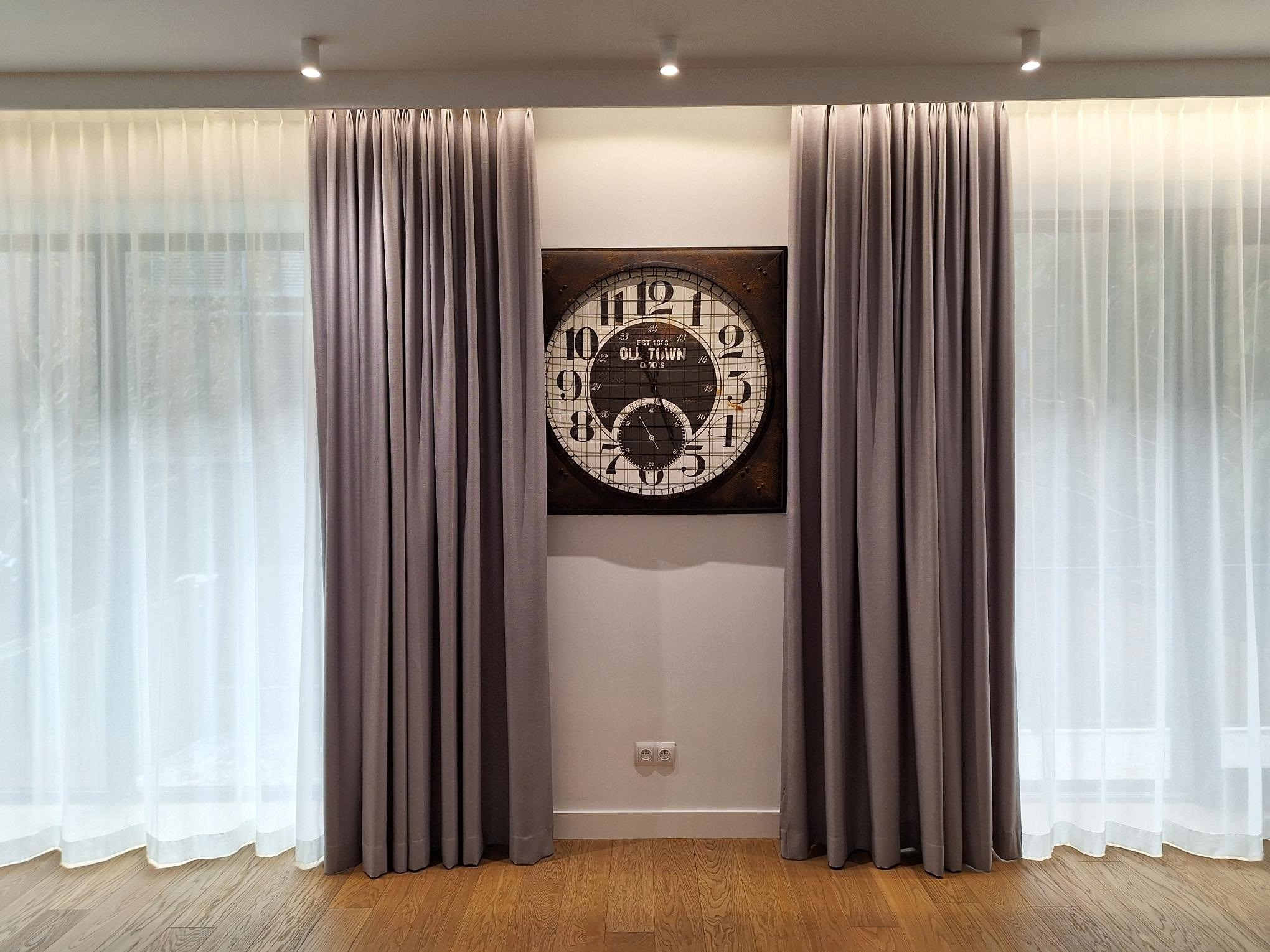 Eleganckie, długie zasłony w odcieniach szarości i białe firany, symetrycznie ułożone po bokach ściany z dekoracyjnym zegarem. Parkiet na podłodze.