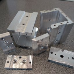 JTH - Precyzyjnie wykonane, srebrne, metalowe elementy z otworami, prawdopodobnie po obróbce skrawaniem, leżące na szarej powierzchni. Widoczne ślady po frezowaniu.