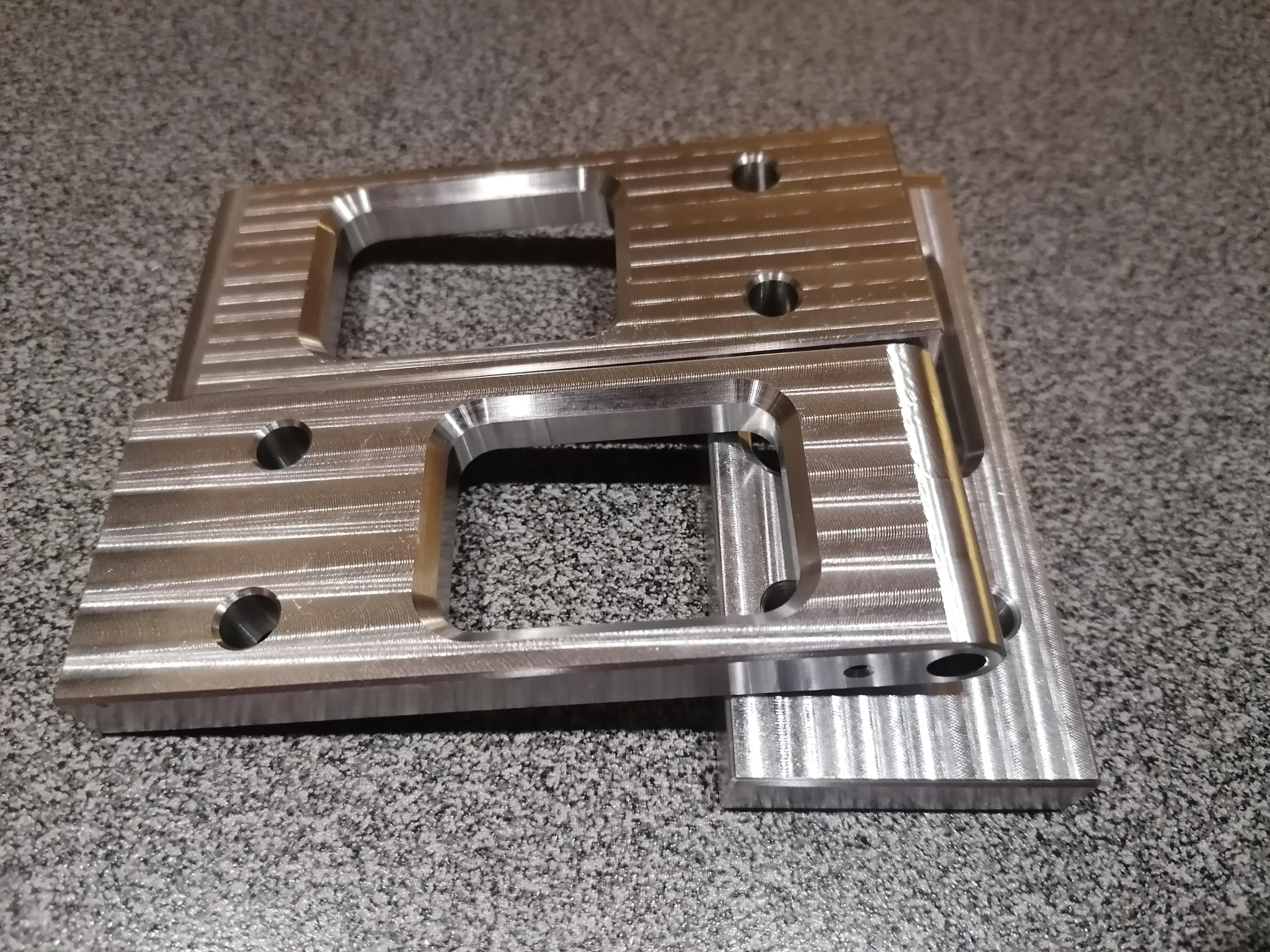 Dwa srebrne, metalowe elementy z otworami i frezowanymi rowkami, ułożone jeden na drugim na szarym, nakrapianym tle. Widoczne ślady po narzędziach.