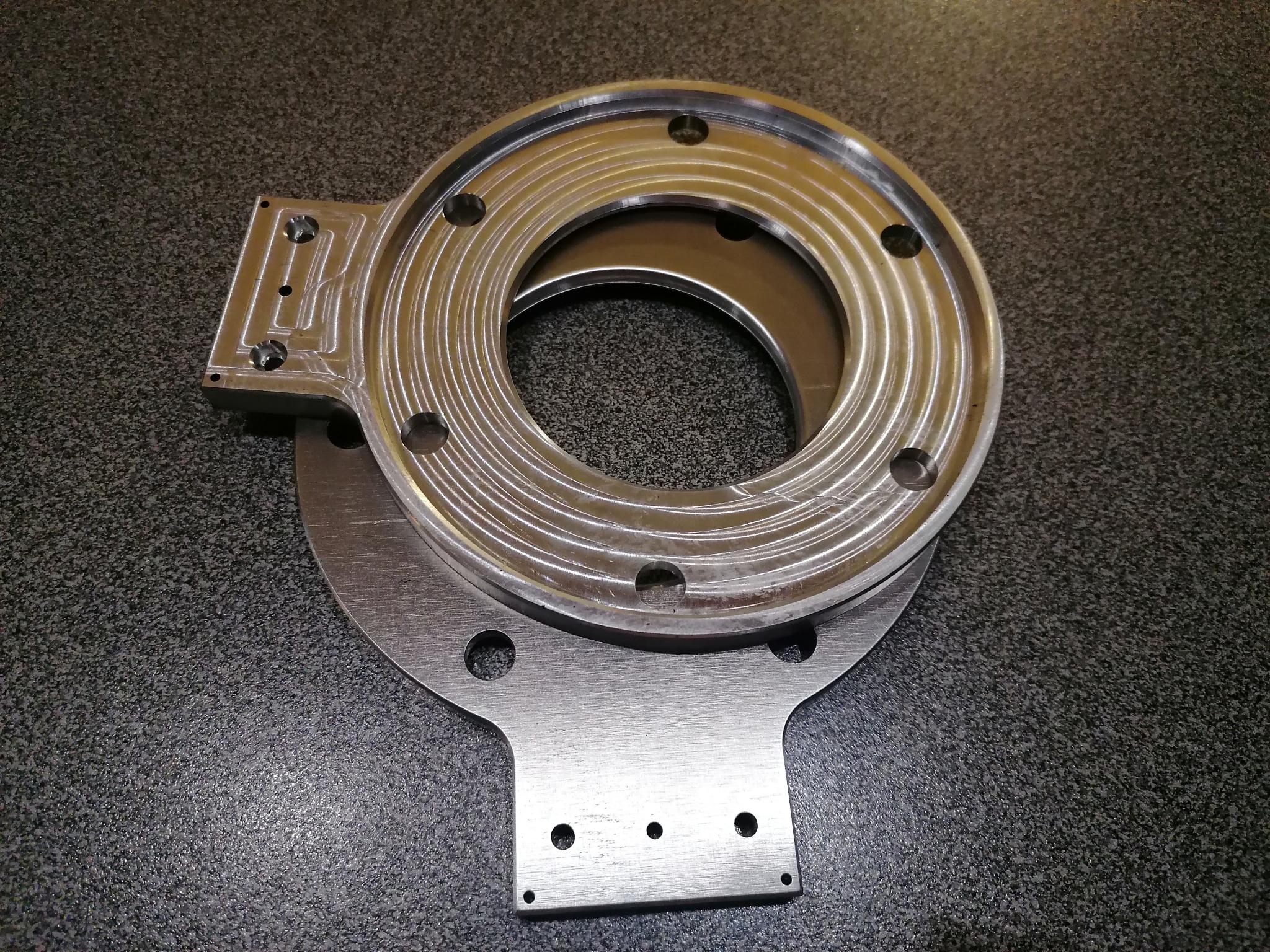 Dwie metalowe części, okrągła i prostokątna, z precyzyjnie wywierconymi otworami i frezowanymi rowkami, leżące na szarej powierzchni. Detale obróbki.