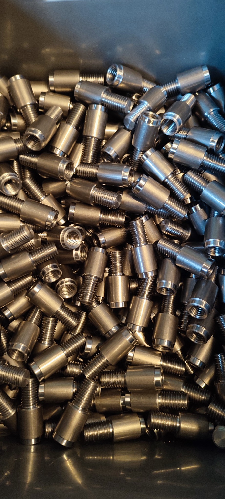 Partia precyzyjnie wykonanych, metalowych śrub z gwintem i cylindrycznym korpusem, ułożonych w pojemniku. Widoczne detale obróbki i metaliczny połysk.