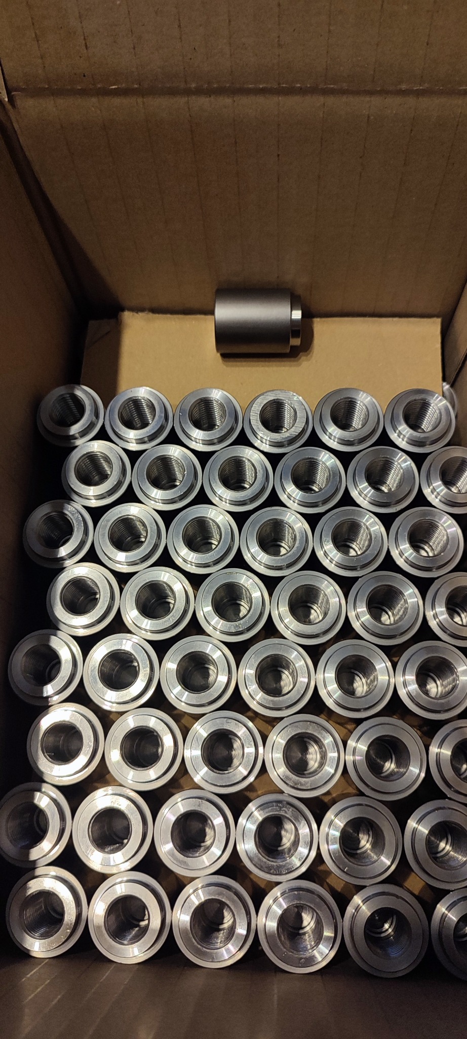 Srebrne, metalowe tuleje z gwintem wewnętrznym ułożone w kartonie, jedna tuleja obok, widok z góry. Precyzyjna produkcja seryjna.