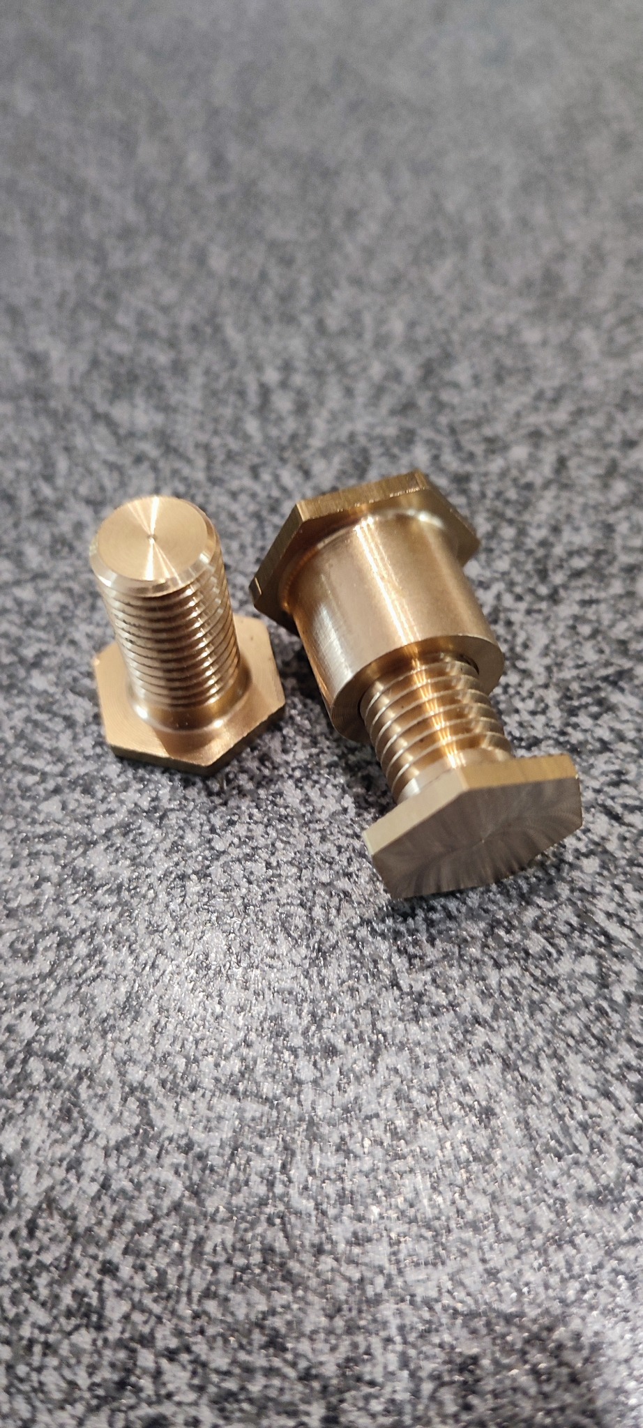 Dwa mosiężne, gwintowane elementy z sześciokątnymi podstawami leżące na szarej, teksturowanej powierzchni. Widoczne ślady po obróbce.