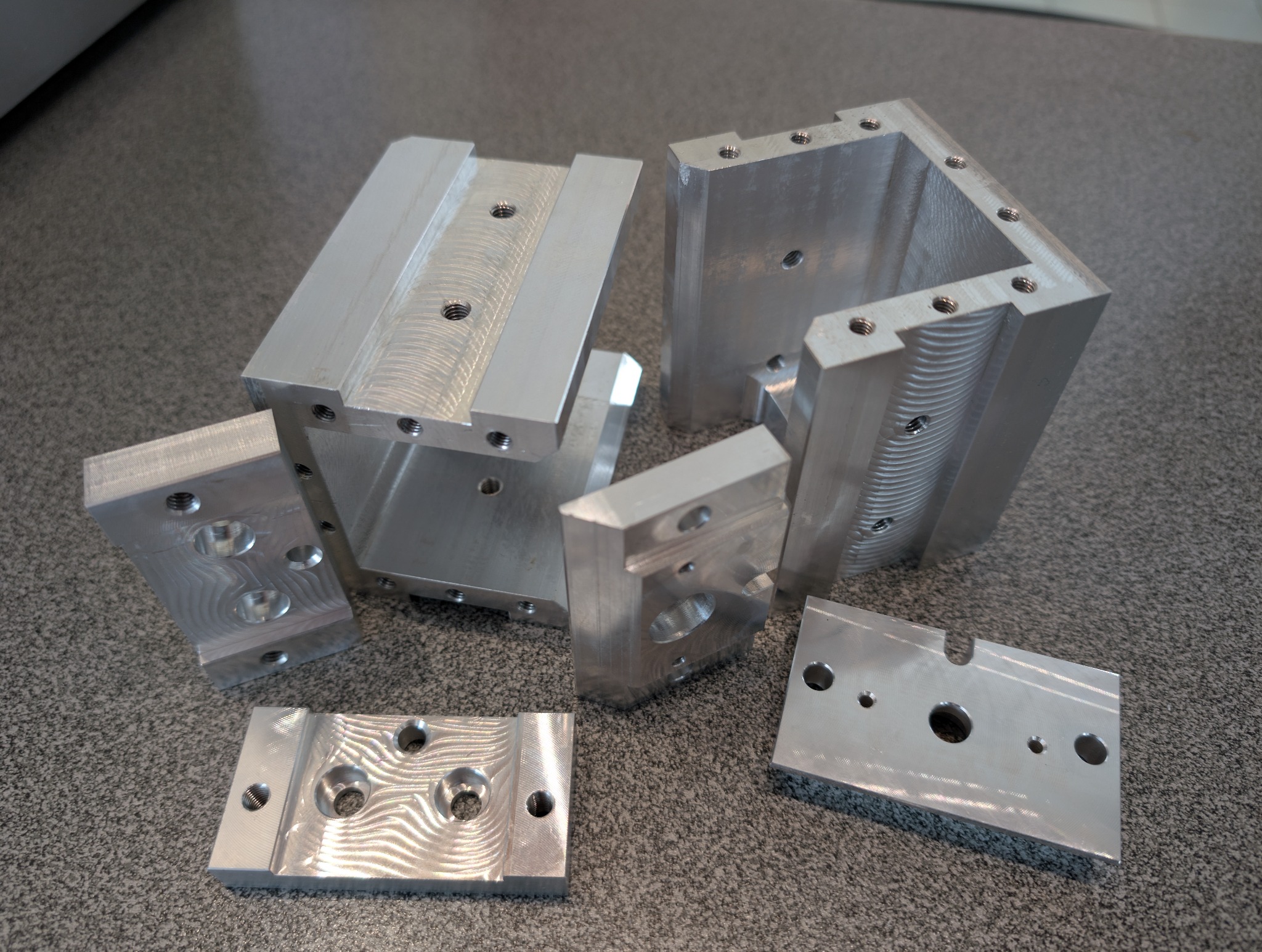 Precyzyjnie wykonane, srebrne, metalowe elementy z otworami, prawdopodobnie po obróbce skrawaniem, leżące na szarej powierzchni. Widoczne ślady po frezowaniu.