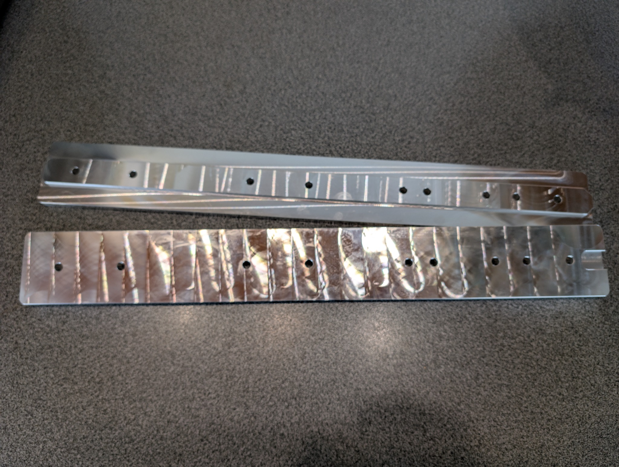 Dwie metalowe listwy z precyzyjnymi otworami i frezowaniami, leżące na szarym tle. Widoczne ślady po narzędziach, sugerujące proces wytwarzania.