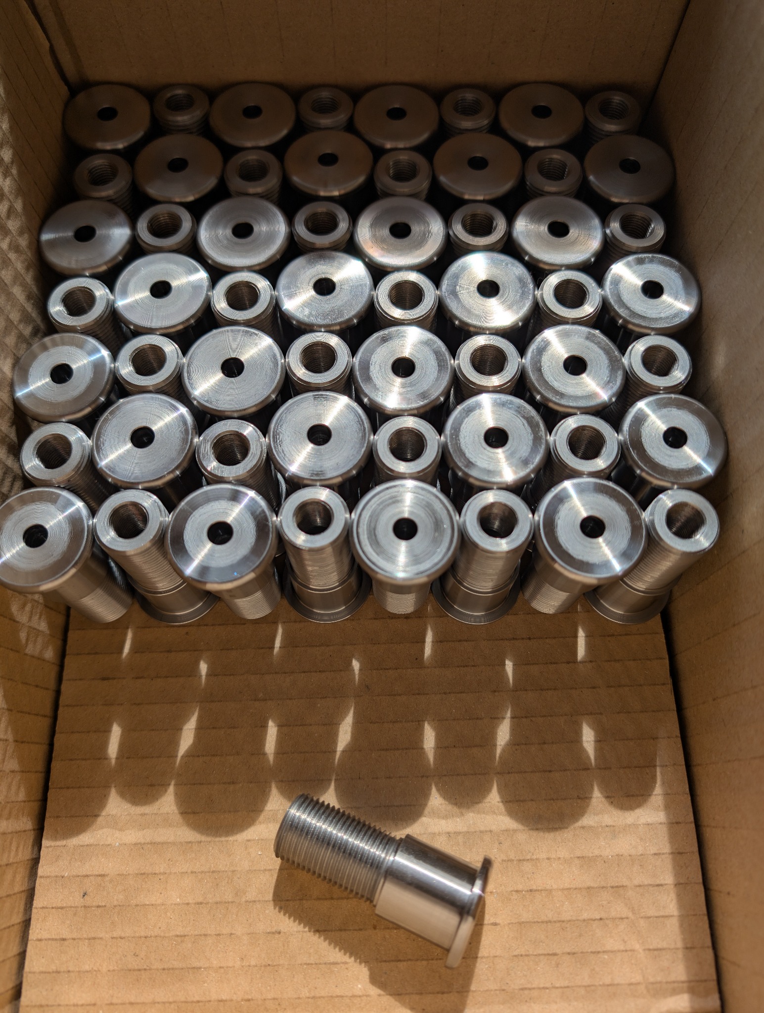 Partia metalowych elementów z gwintem, ułożonych w kartonowym pudełku. Jeden element leży poza szeregiem, ukazując detale gwintu i obróbki.